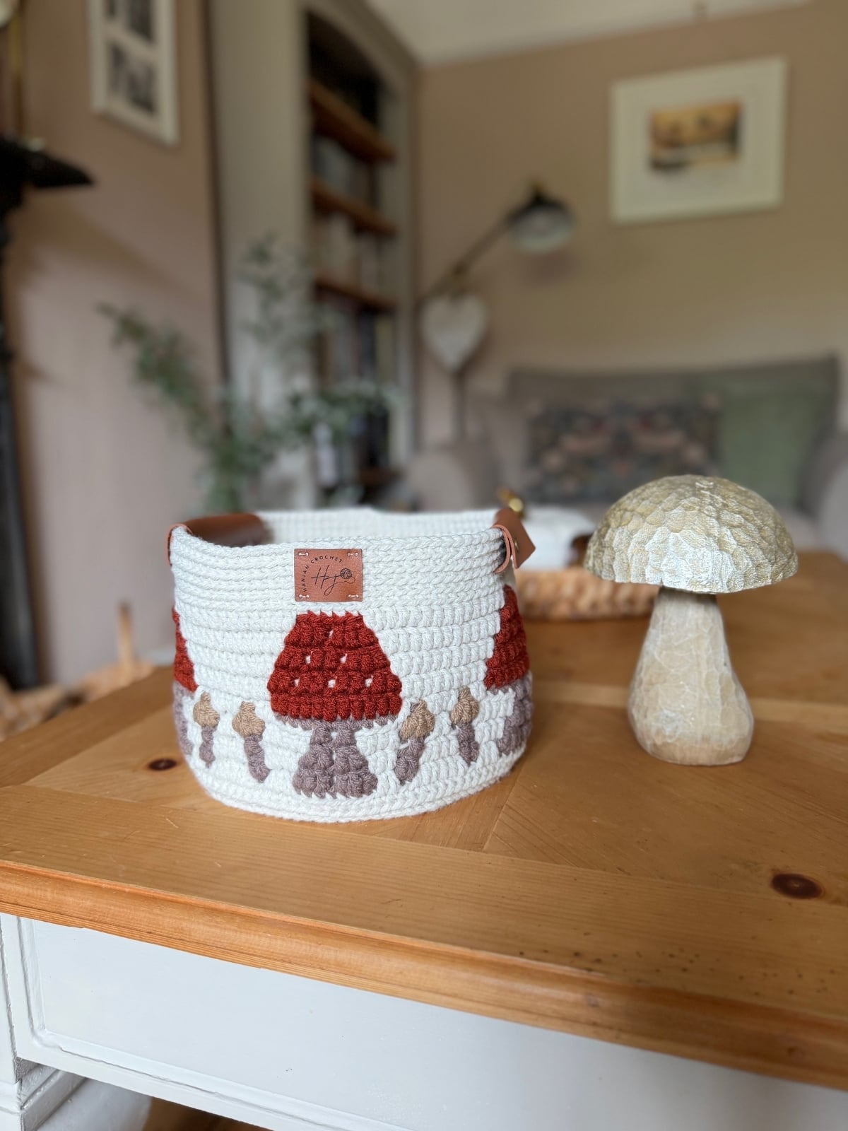 A crocheted basket with a charming mushroom pattern, made using a crochet storage basket pattern, sits on a wooden table next to a decorative stone mushroom in a cozy, softly lit living room.