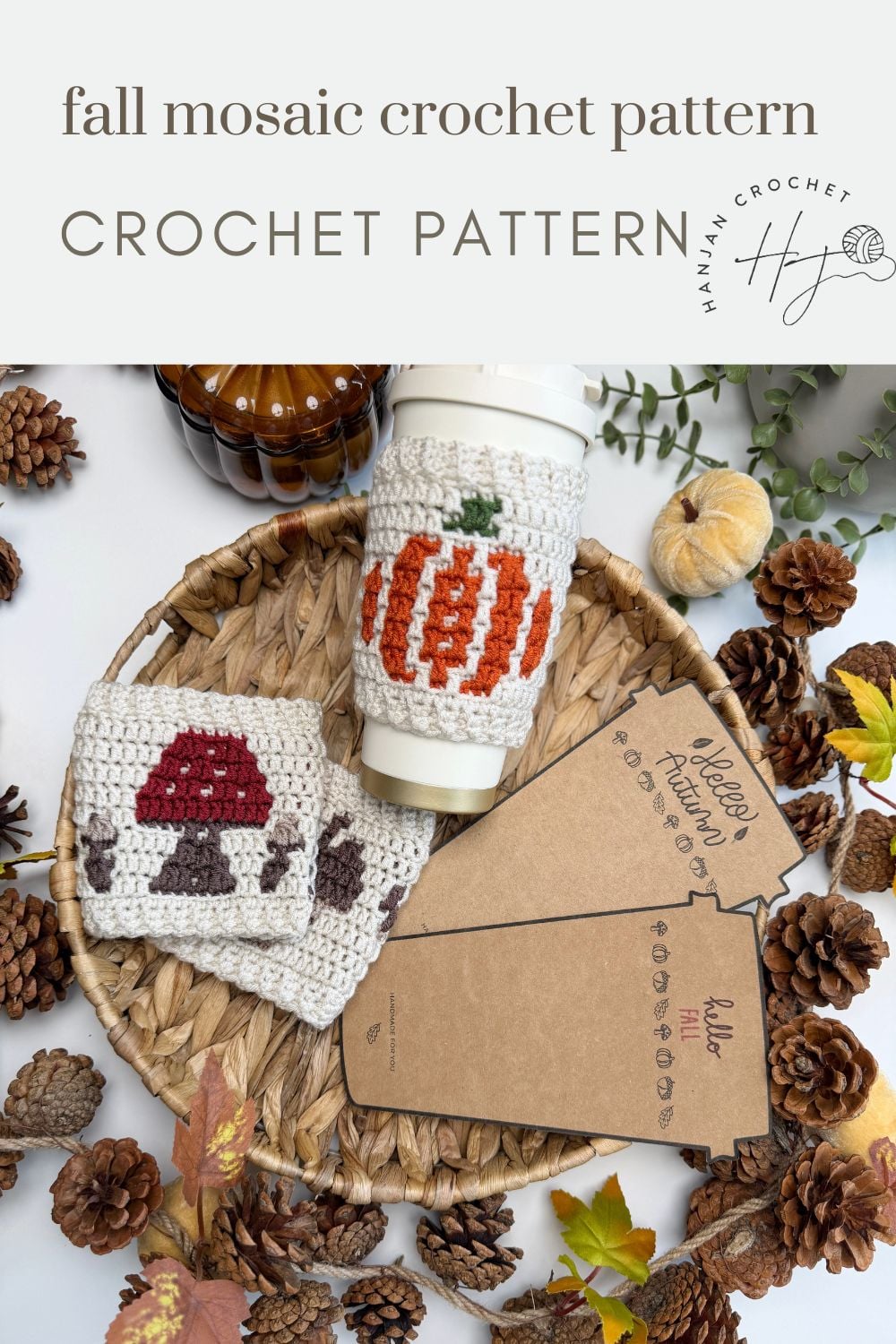 Woven basket with crochet cup sleeves featuring pumpkin and mushroom designs, crafted using a charming crochet cup holder pattern, surrounded by pinecones, autumn leaves, and notecards on a white surface.