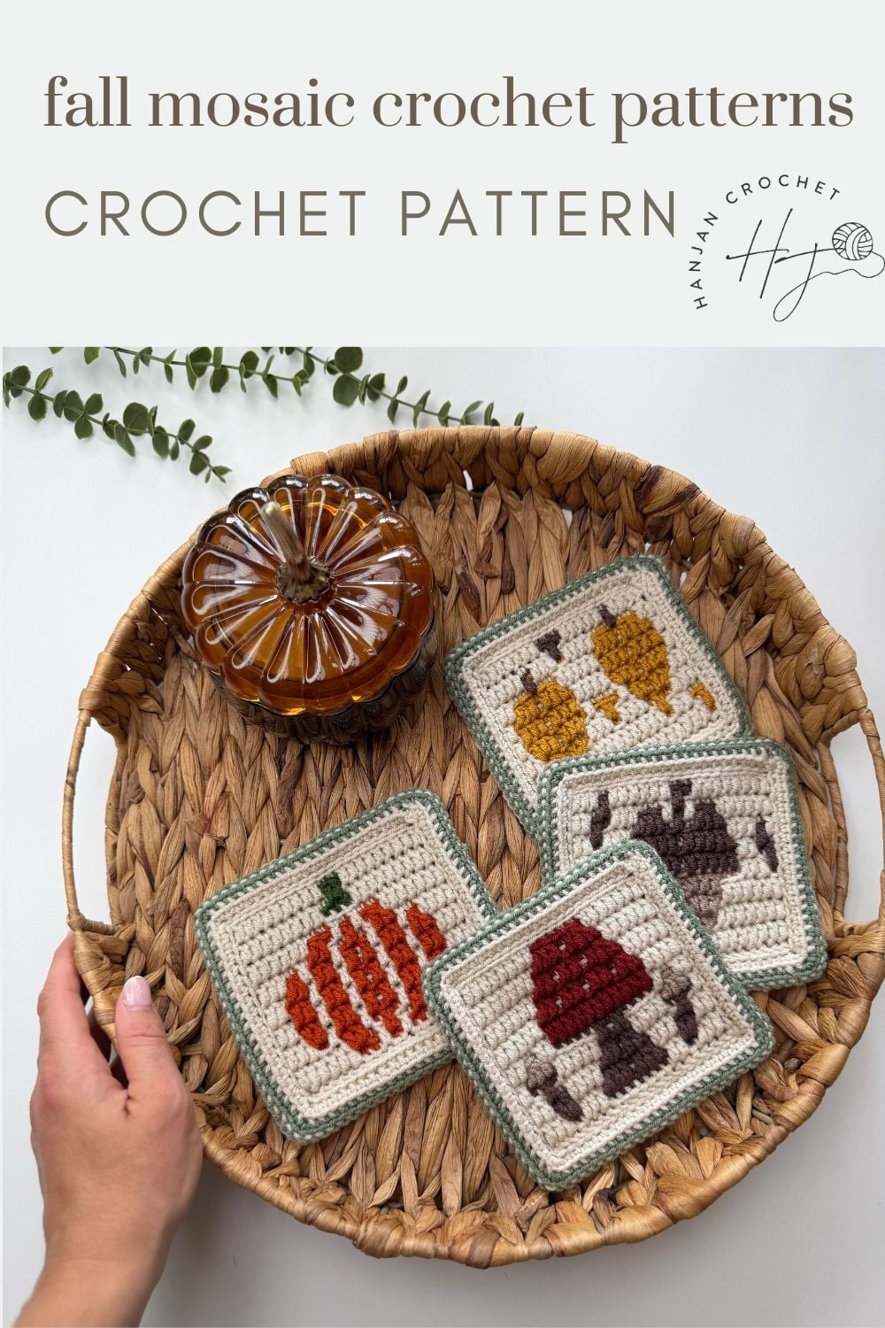 A hand holds a wicker basket with a glass pumpkin and four crochet coasters featuring autumn designs: pumpkin, mushrooms, acorns, and leaves.