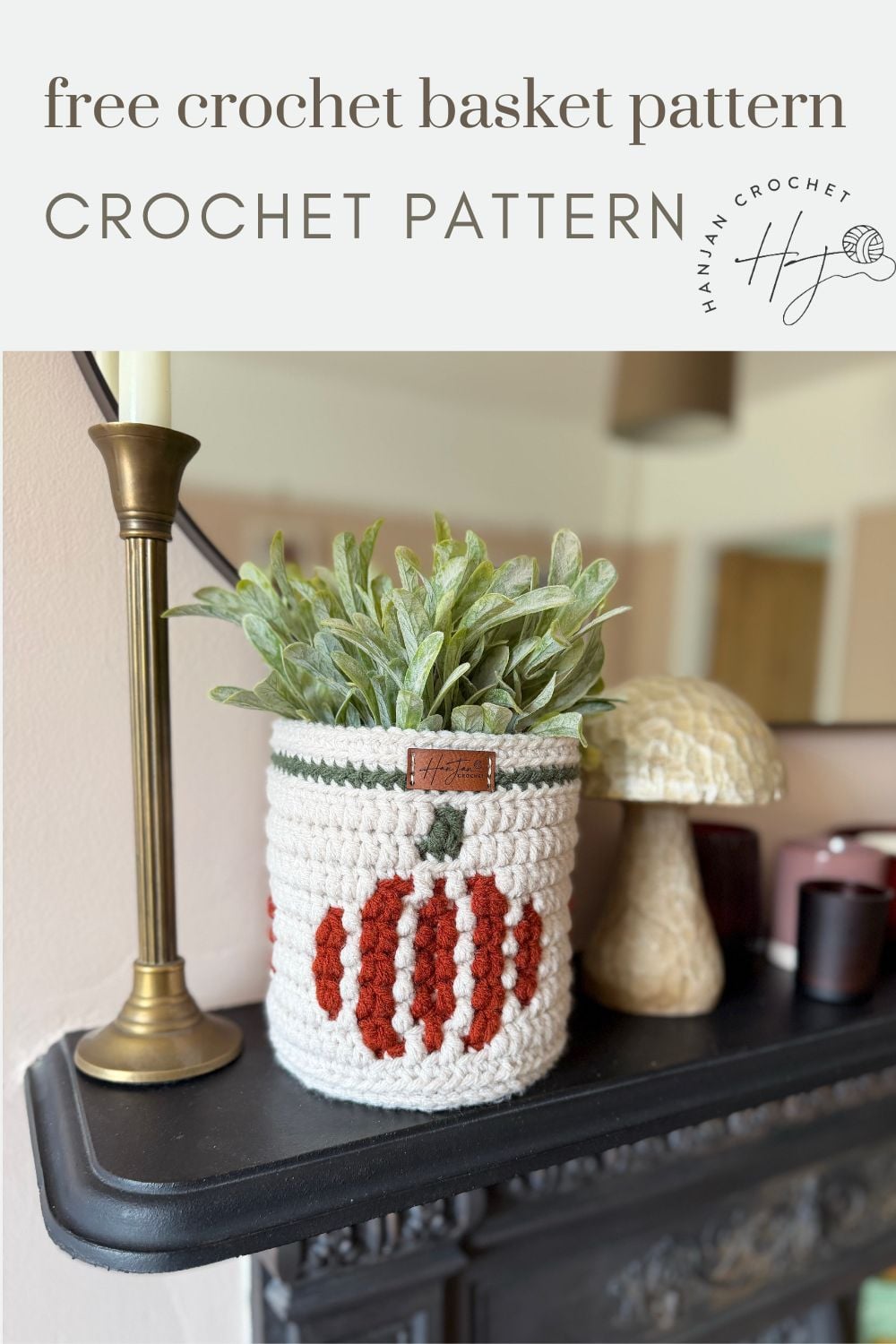 A crochet pumpkin basket with red and white stripes holds a green plant on a black mantel, next to a candle, ceramic mushroom, and pink cup.
