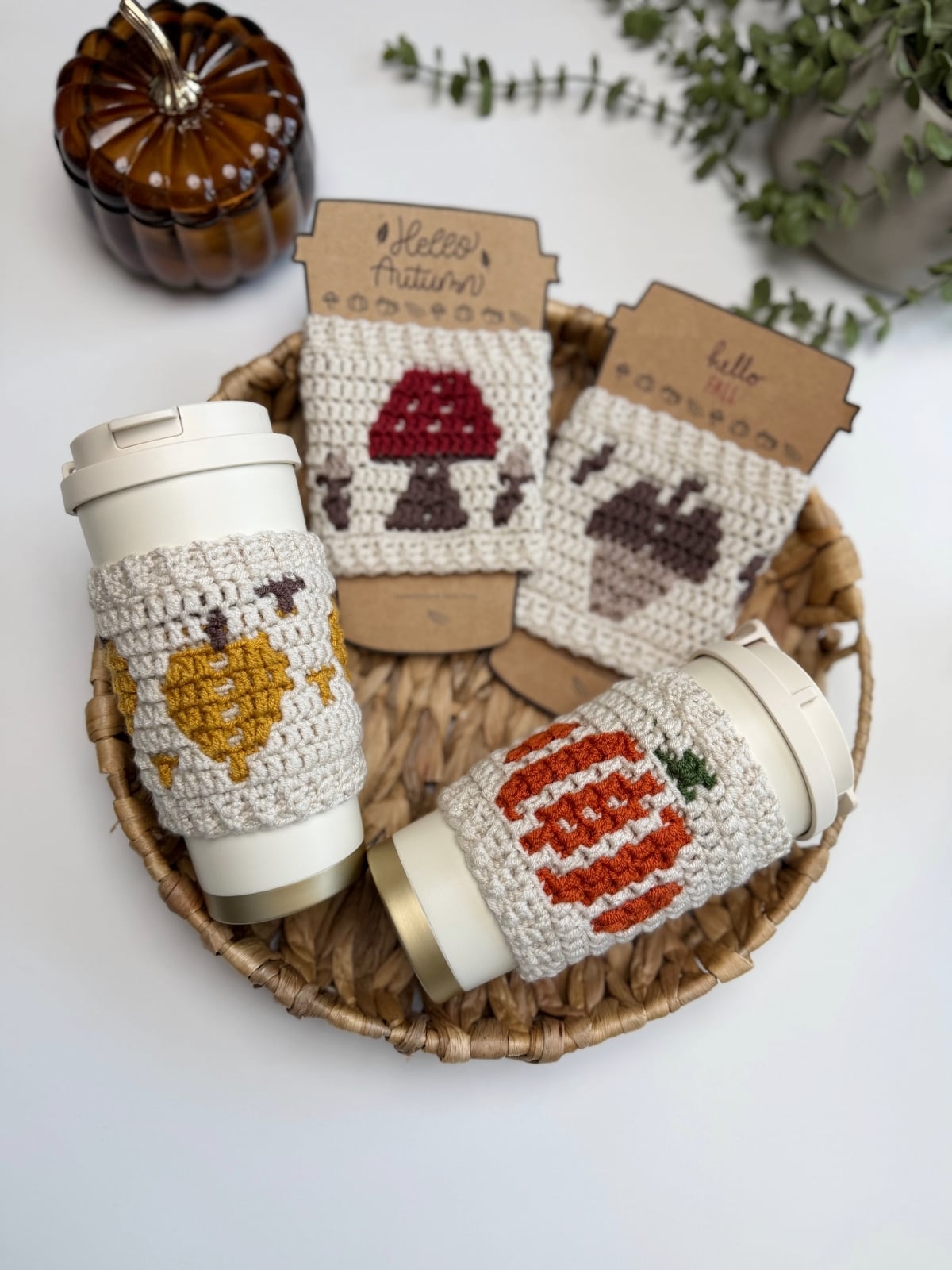 Two reusable cups with crocheted autumn-themed sleeves in a wicker basket, showcasing an elegant crochet cup holder pattern, with two additional crochet sleeves featuring a mushroom and acorn displayed upright behind them.