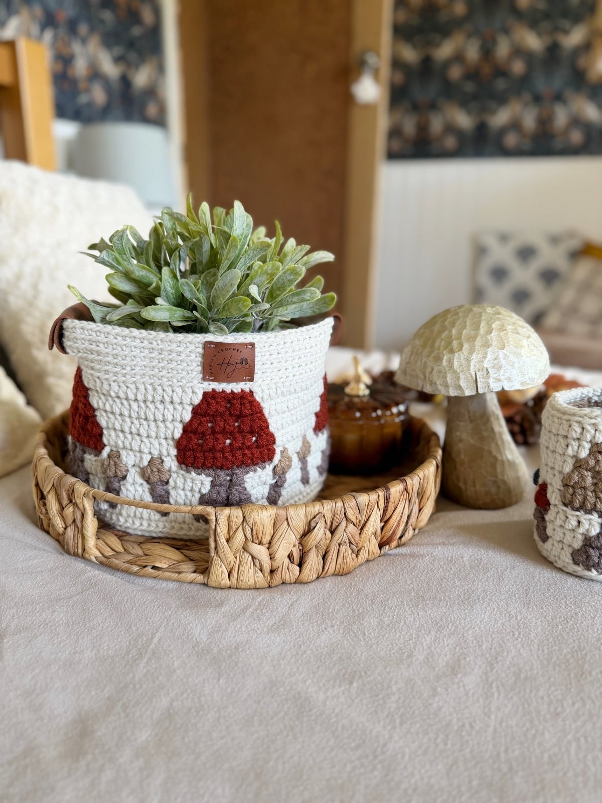 A crocheted basket with a plant sits on a woven tray, next to a crochet cup and a decorative mushroom on a beige surface, showcasing the charm of a unique crochet storage basket pattern.