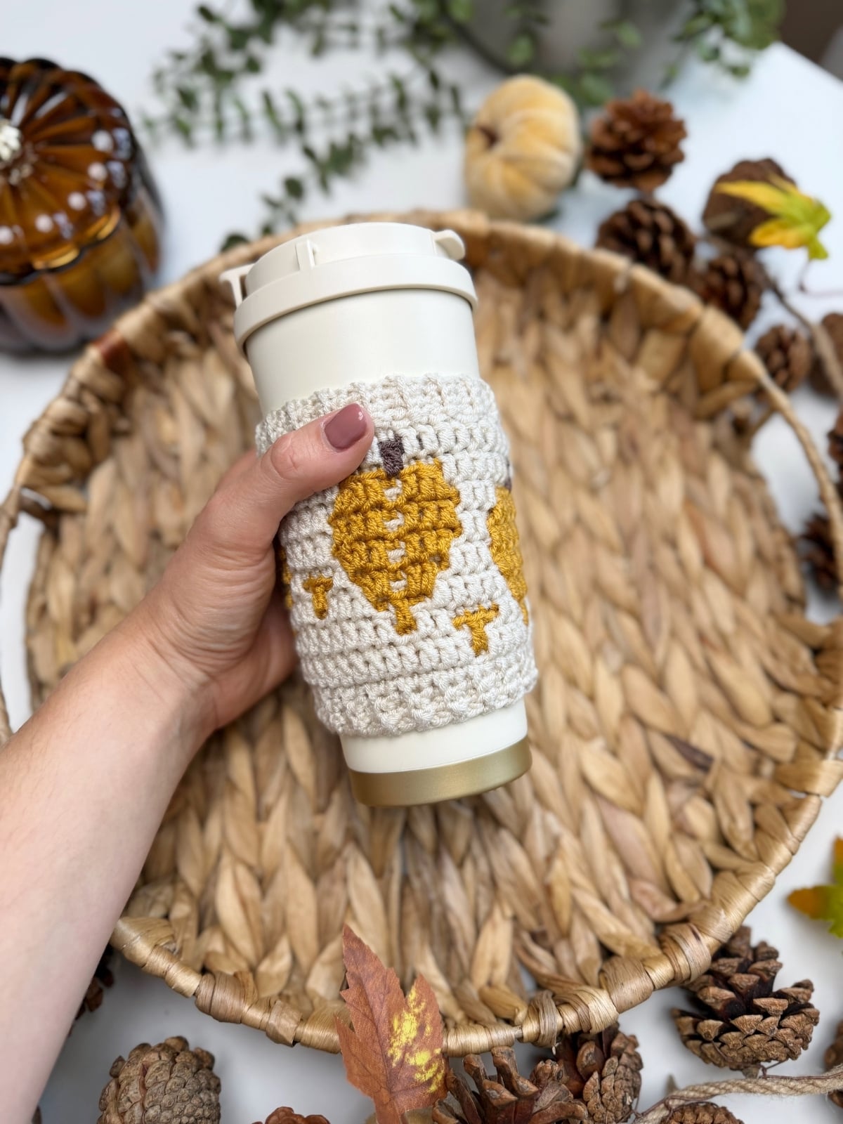 A hand holds a reusable cup with a crocheted sleeve featuring yellow leaf designs, made using a crochet cup holder pattern, above a woven tray with pine cones and autumn decorations nearby.
