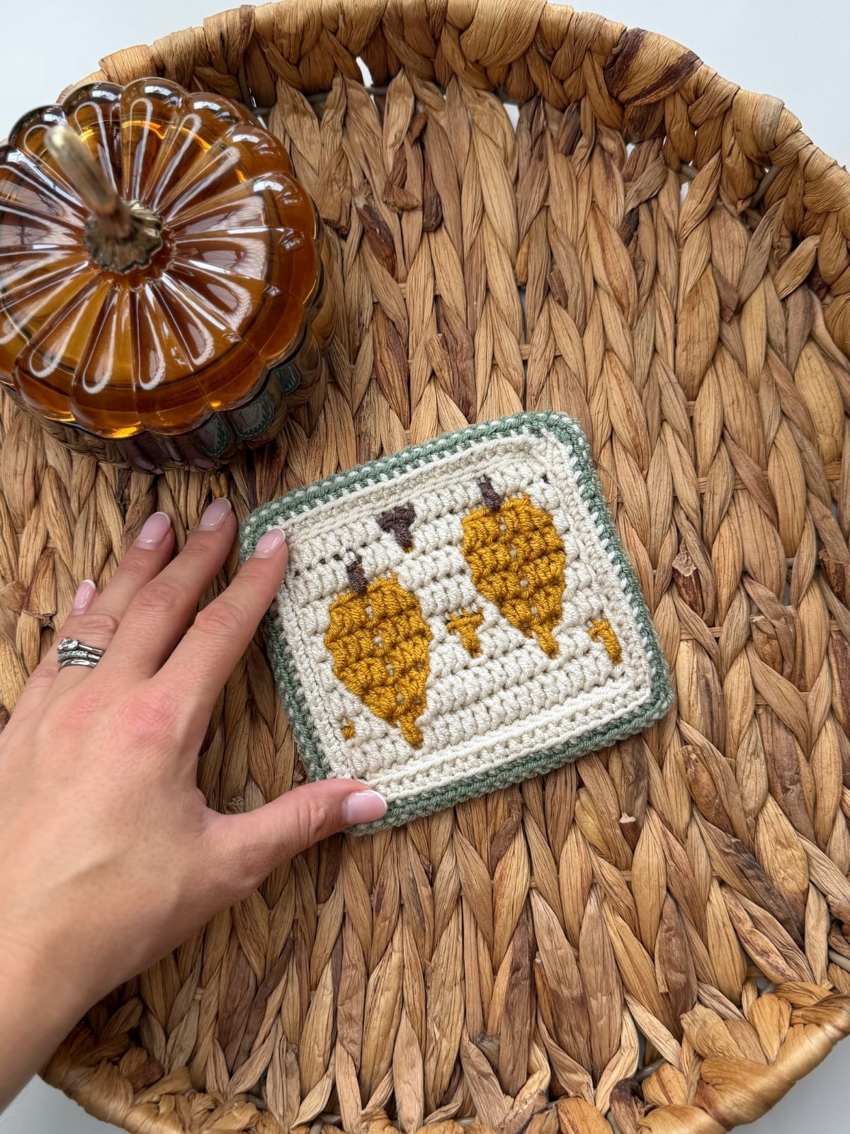 A hand holds a crocheted coaster with yellow leaves, placed in a woven basket next to a glass pumpkin.