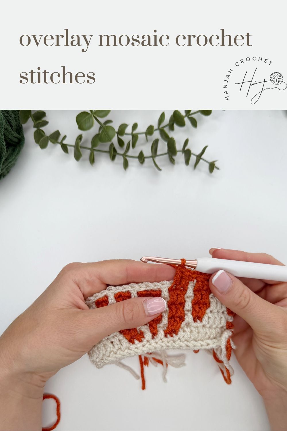 Two hands crochet a white and orange mosaic pattern with a metal hook, with green yarn and a sprig of greenery on a white surface. Text reads “overlay mosaic crochet stitches.”.