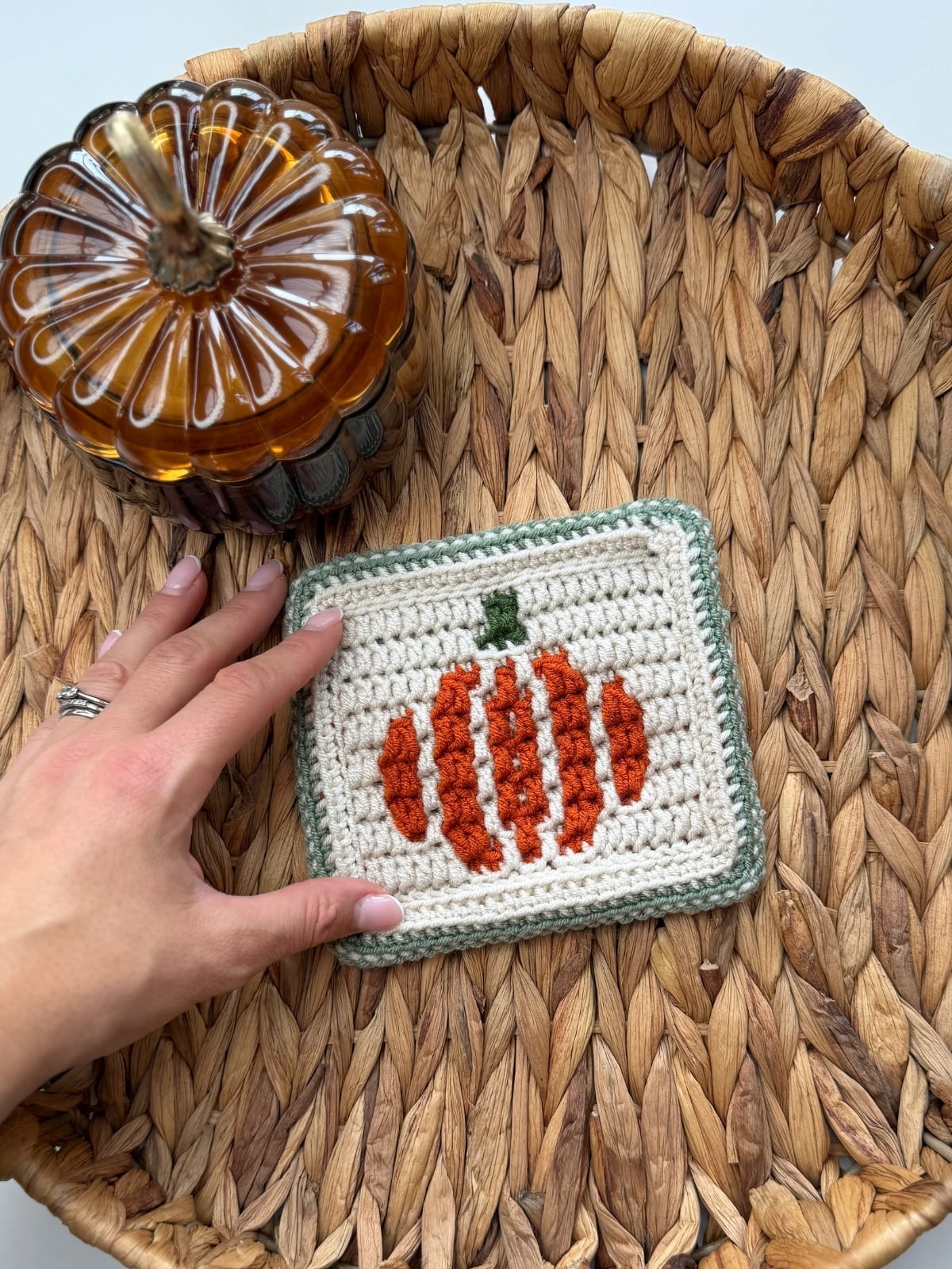 A hand holds a crocheted square with an orange pumpkin design on a woven tray next to a glass pumpkin-shaped jar.