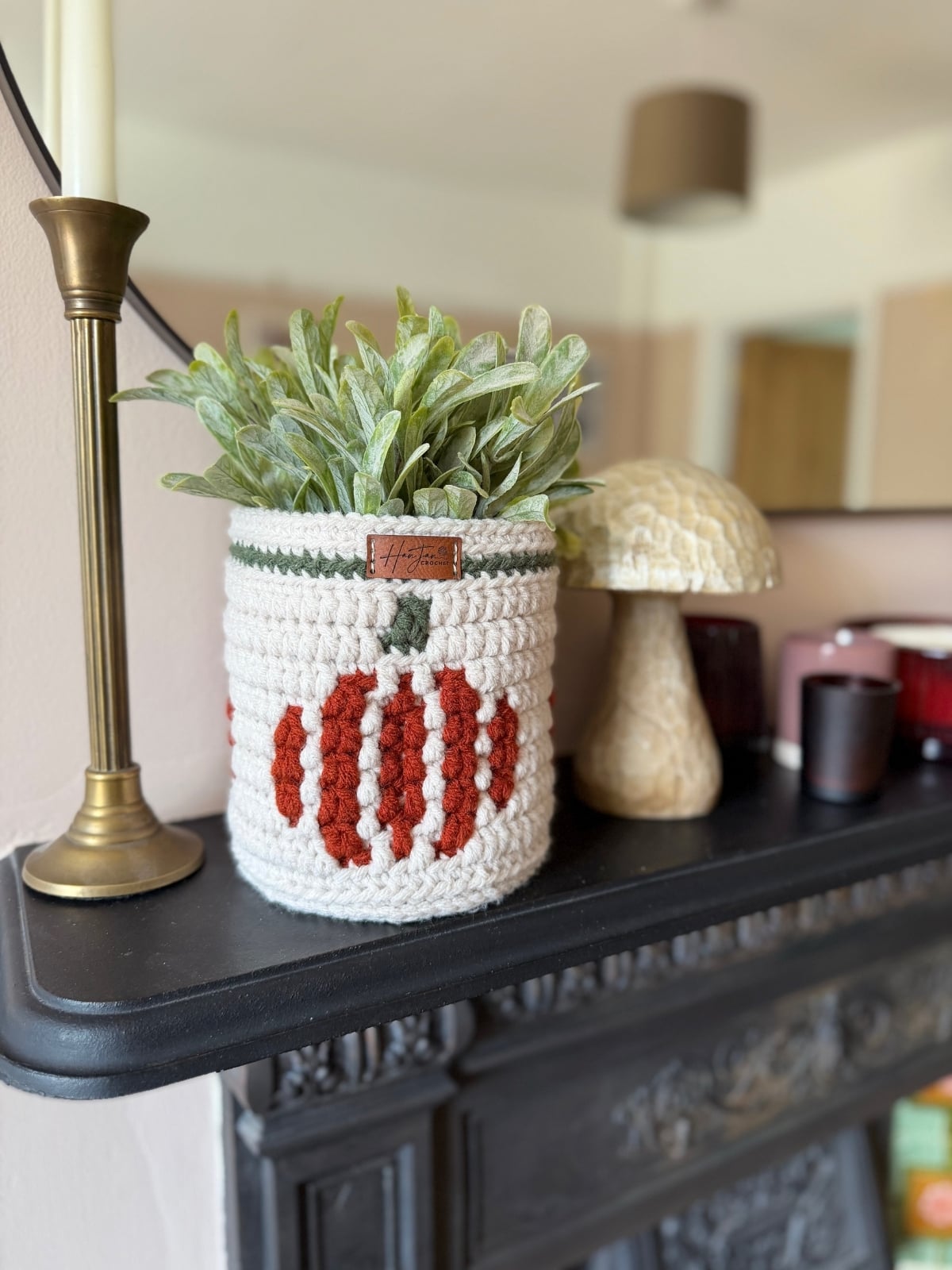 A green plant in a white crocheted pot with red oval patterns sits on a black mantel next to a tall candle, a mushroom ornament, a crochet pumpkin basket, and small decorative items.
