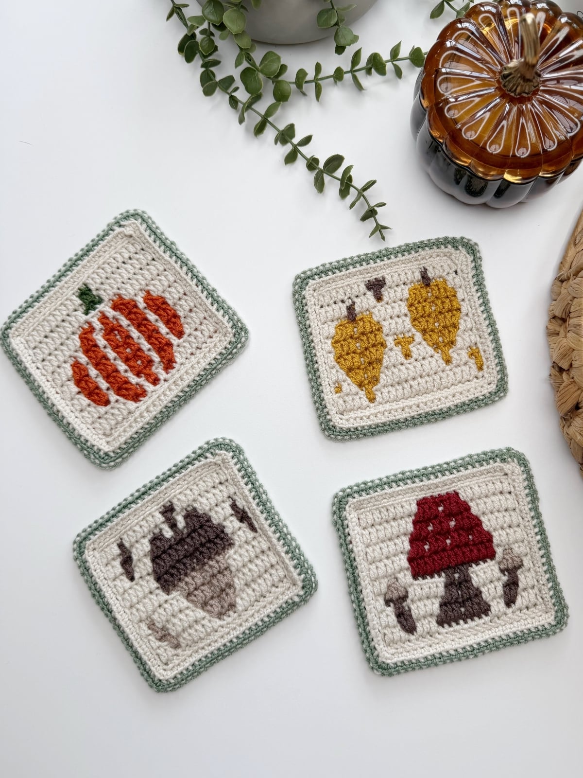 Four crocheted coasters with designs of a pumpkin, two yellow leaves, an acorn, and a red and brown mushroom are arranged on a white surface near plants and a glass pumpkin.