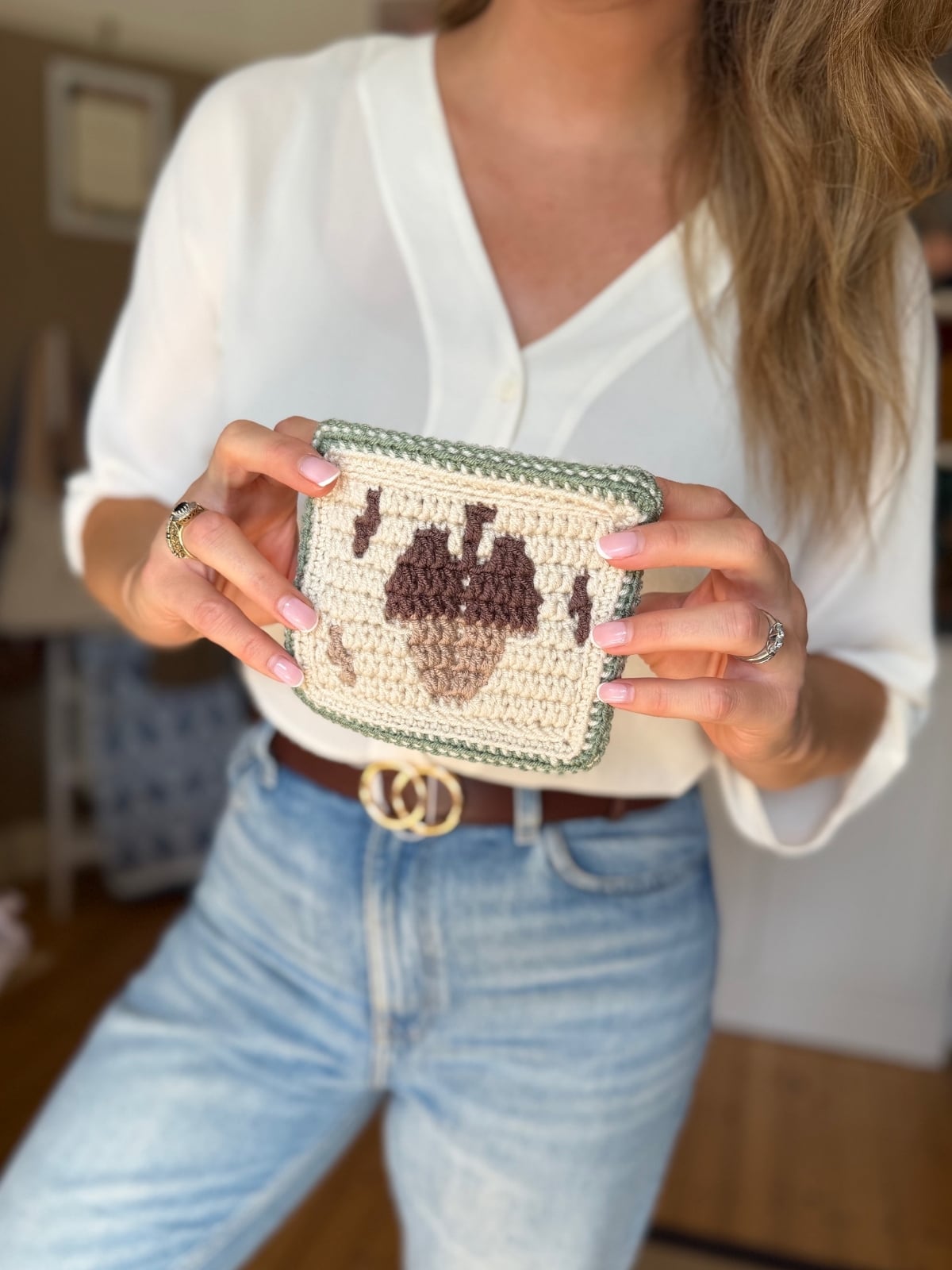 A woman in a white blouse and jeans holds a crocheted piece featuring an acorn design, proudly displaying her intricate acorn crochet pattern.