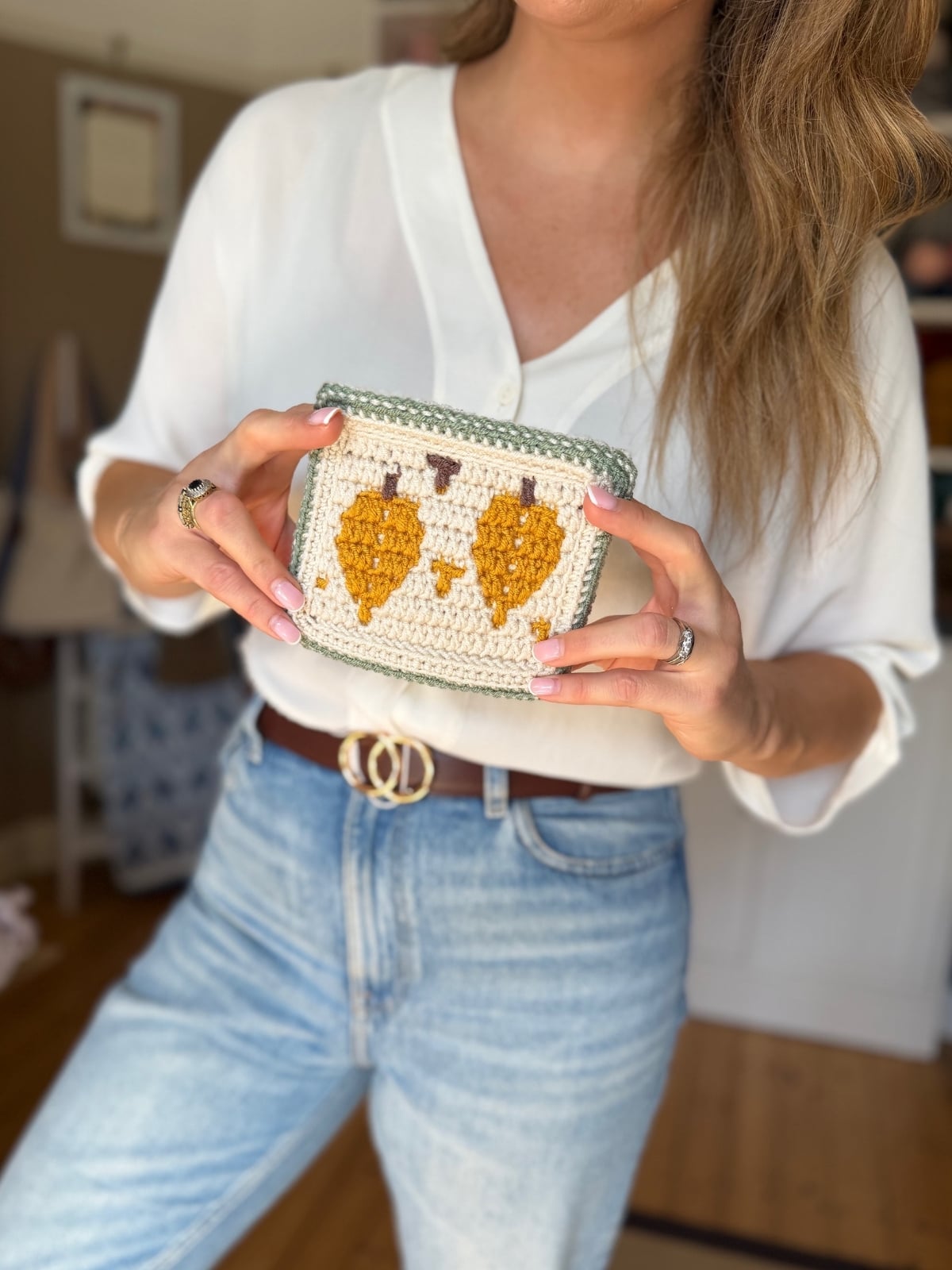 A person in a white shirt and jeans holds a small rectangular crocheted pouch featuring a charming fall leaf crochet pattern with two yellow leaves.