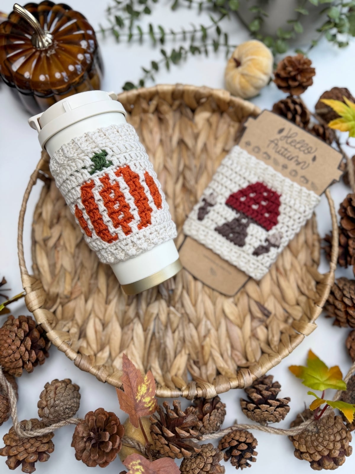 A reusable cup with a crocheted pumpkin sleeve and a packaged crochet mushroom sleeve—crafted using a charming crochet cup holder pattern—sit on a woven tray surrounded by pinecones, leaves, and autumn decorations.