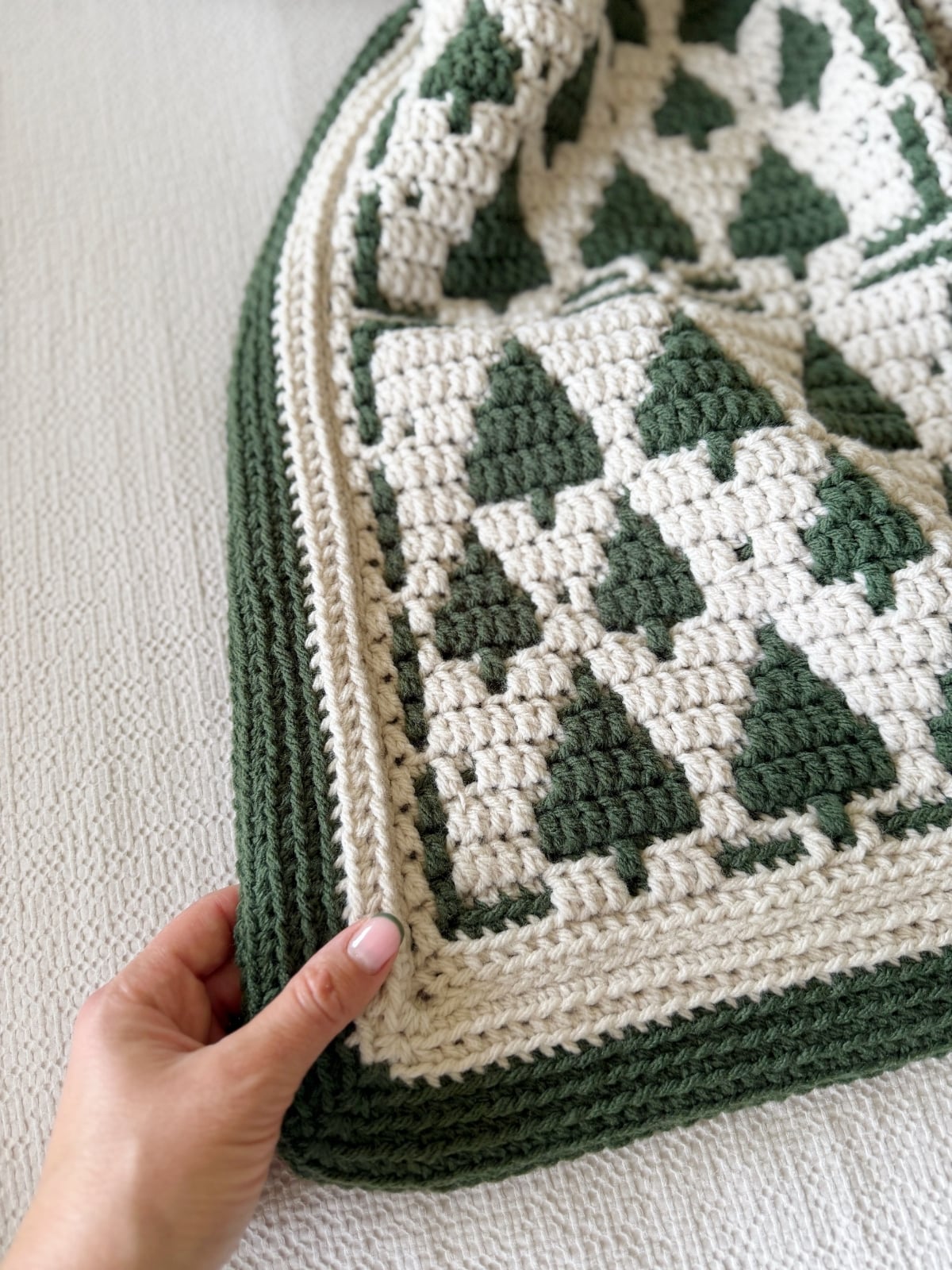 A hand holds the corner of a crocheted blanket featuring a pattern of dark green pine trees on a cream background with a ribbed envelope border.