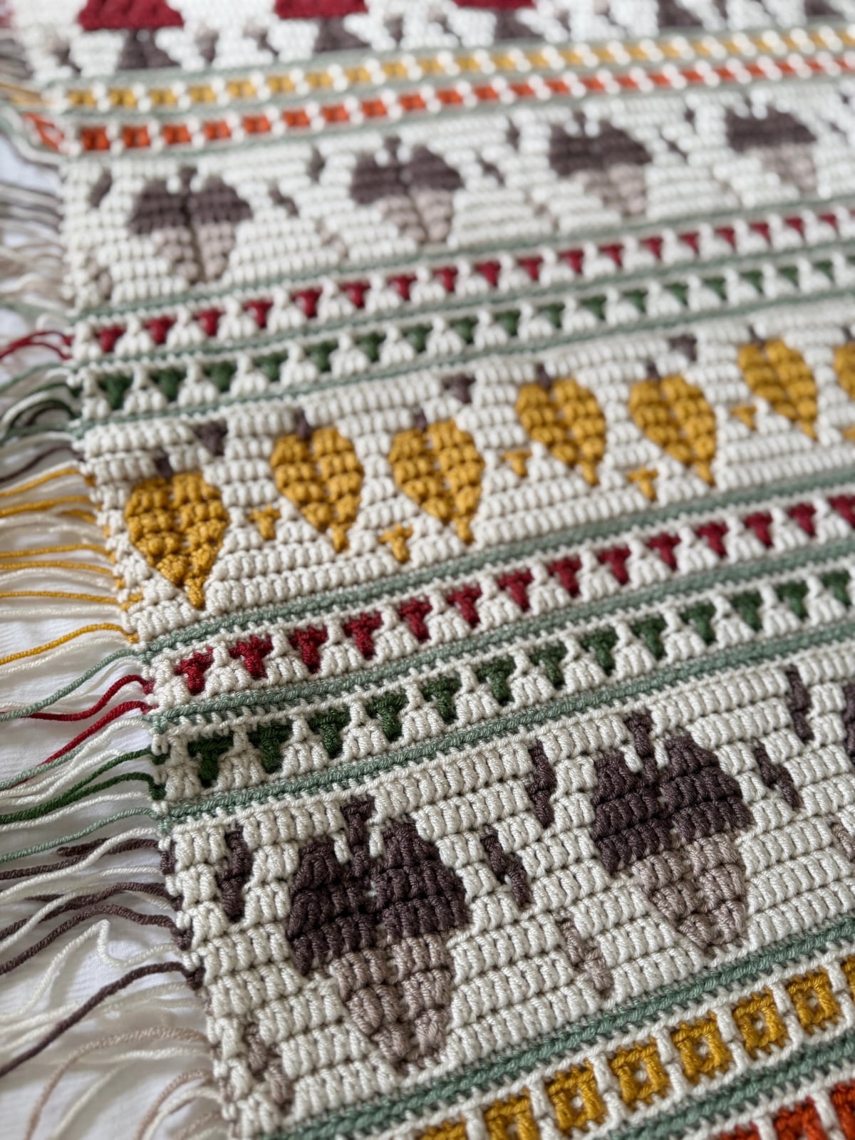 A crocheted tapestry featuring rows of acorns, leaves, and geometric patterns in cream, brown, gold, green, and red, finished with a crochet envelope border and fringed edges.